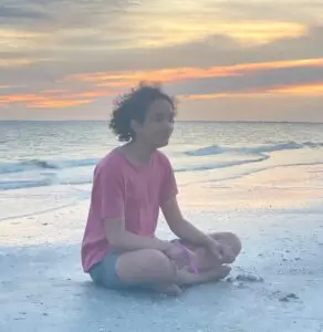 A young man sit cross legged on a sandy beach