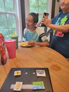 A young man is smiling and eating a meal at a table. He has a picture communication book with him.