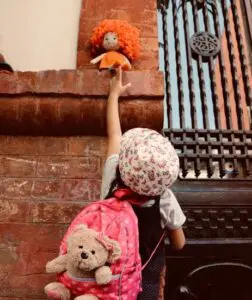 A young girl is reaching up towards a doll that is on a ledge above her.