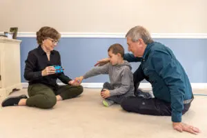 A young boy is sitting on the floor, reaching to exchange a picture to communicate to a woman who is holding a toy train.