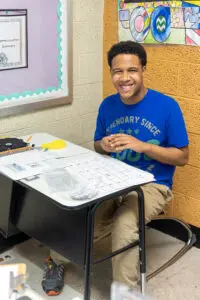 A smiling high school aged young man is sitting at his desk in a classroom.