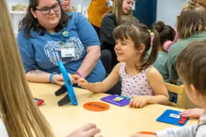 A young girl is smiling and using a speech generating device in a classroom